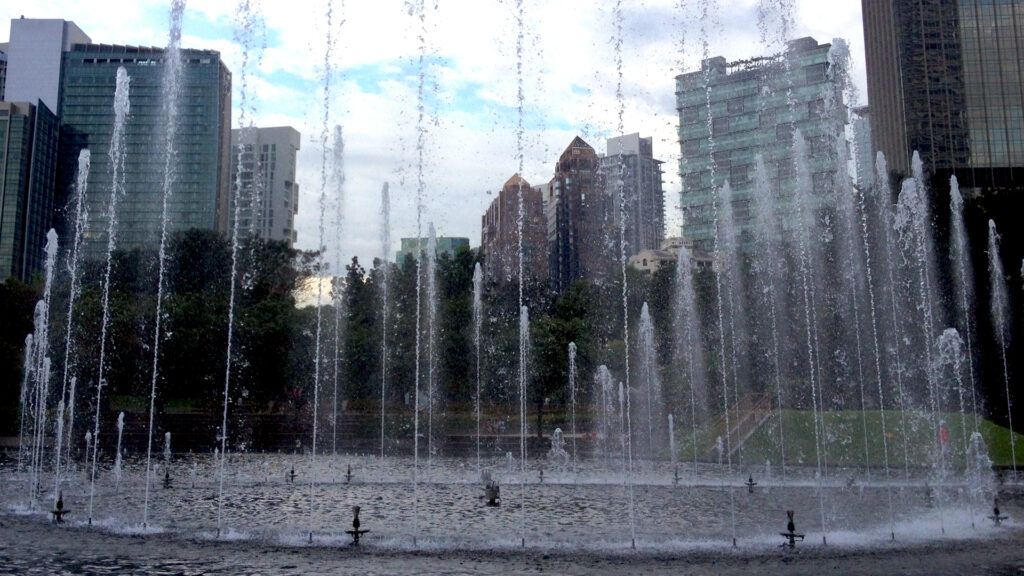 Skyscrapers line the streets behind a spouting fountain